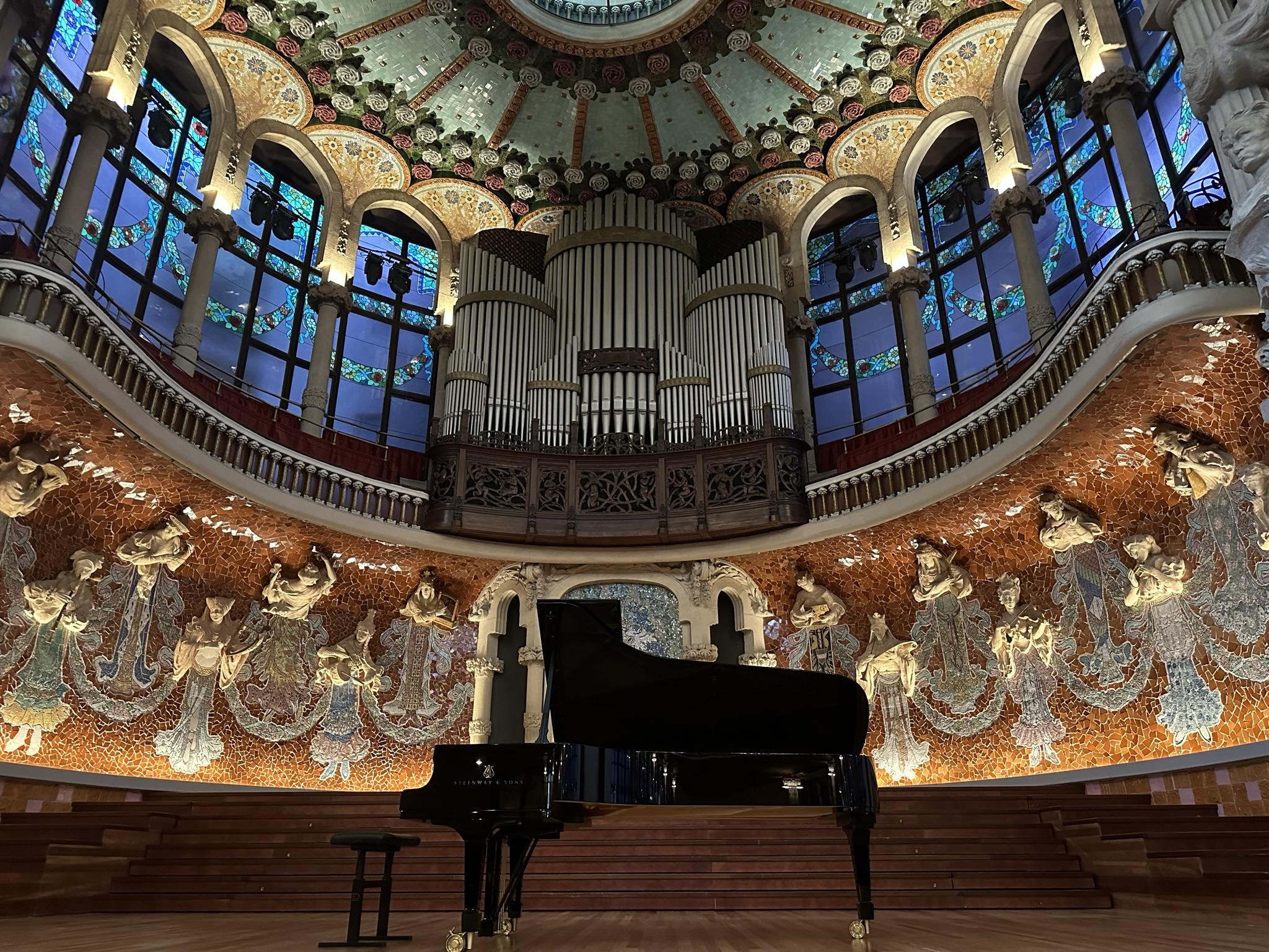 Stage of Palau musica. A piano is at the stage center. Colorful musiac is in the background with lifelike status coming out of the walls of the stage. Each statue holding a different instrument.